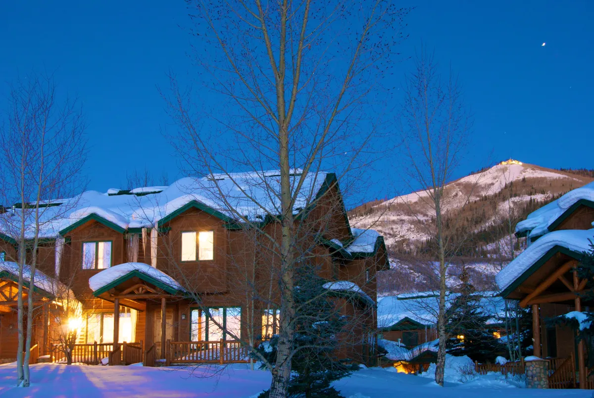 A winter exterior view of the property, featuring snow-covered rooftops and warmly lit windows set against a dramatic mountain backdrop. The scene captures a tranquil, inviting atmosphere with clear hints of the surrounding ski terrain.