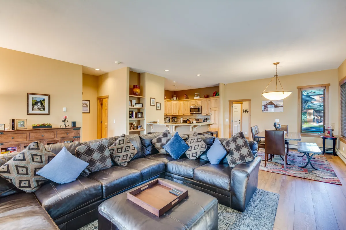 A wide-angle view of the open-concept interior, capturing a large sectional sofa, a wooden dining table set for meals, and the adjoining kitchen area. The room feels airy and filled with natural light, highlighting the seamless flow between living, dining, and kitchen spaces.