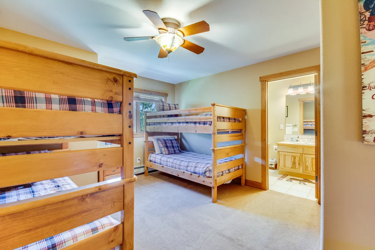 A bright, cheerful bunk room featuring two sets of twin-over-twin bunk beds, a ceiling fan, a window with sunlight filtering in, and a visible bathroom door. The playful layout is perfect for kids or friends to share late-night stories.