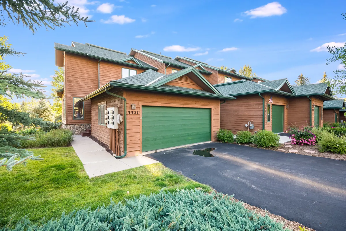 This exterior shot features the home’s wooden facade with a distinctive green garage door, bordered by lush landscaping and a spacious driveway. The well-kept garden and inviting entrance set an active, welcoming tone, and the visible garage subtly hints at practical storage for bikes and gear.