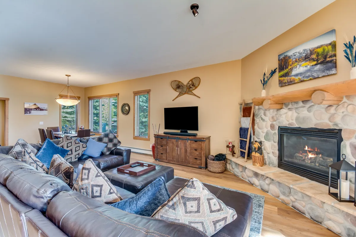 A welcoming living area featuring a cozy fireplace, a comfortable sofa arrangement, tasteful wall art, and a partial view of the dining space. Large windows invite natural light, emphasizing the relaxed and sociable atmosphere. The overall mood is both warm and inviting, ideal for unwinding after a day outdoors or enjoying quiet time indoors.