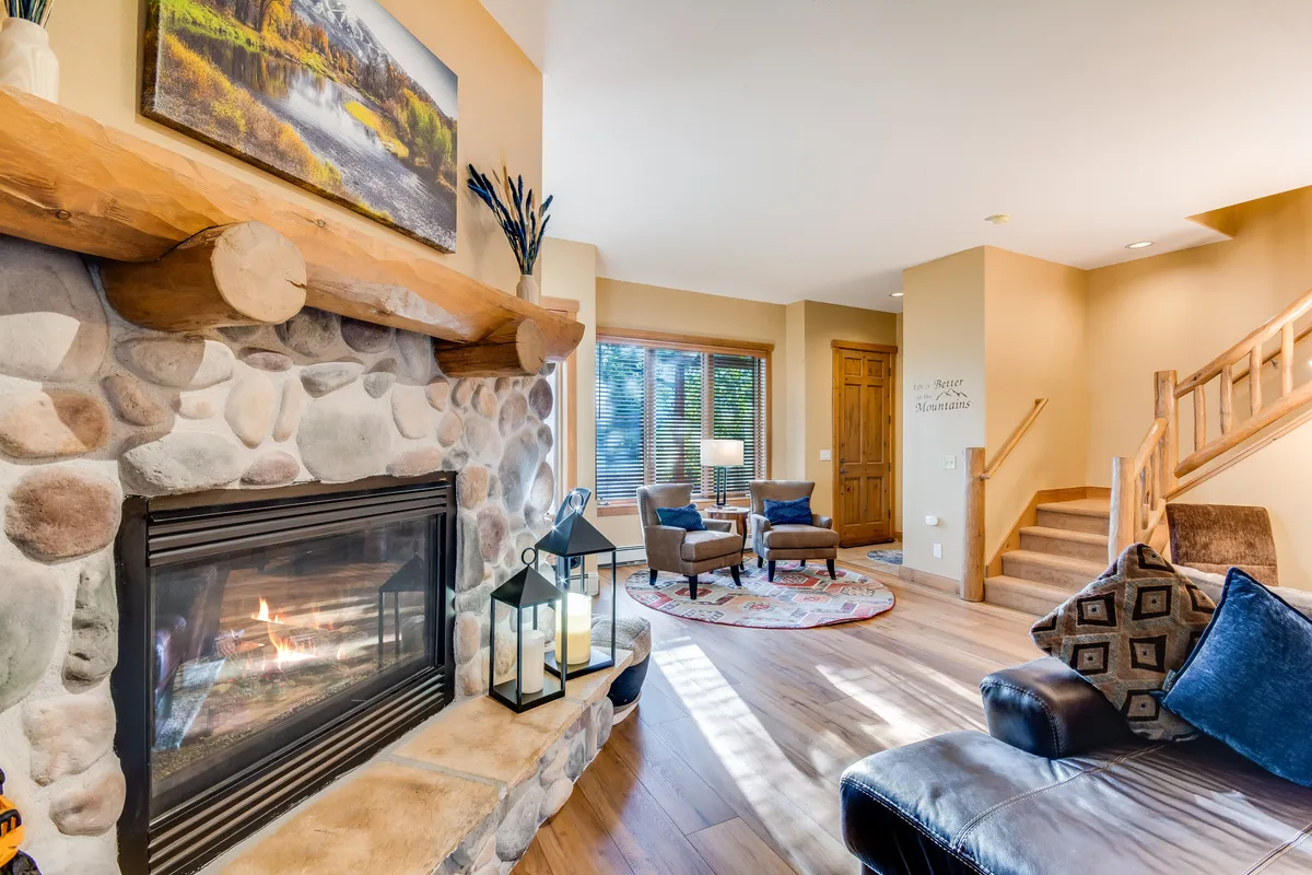 A cozy living room interior featuring a stone fireplace, comfortable seating, wooden staircase, and framed artwork. The scene radiates warmth and comfort—ideal for gathering after outdoor adventures.