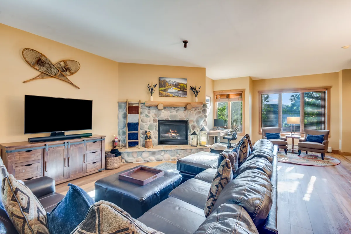 The image showcases a cozy, mountain-inspired living room with a striking stone fireplace as the centerpiece, a plush sectional sofa perfect for gathering, tasteful wooden furniture, and warm lighting. Decorative wall art adds personality, and the setup is ideal for relaxing, entertaining, or warming up after a day on the slopes.