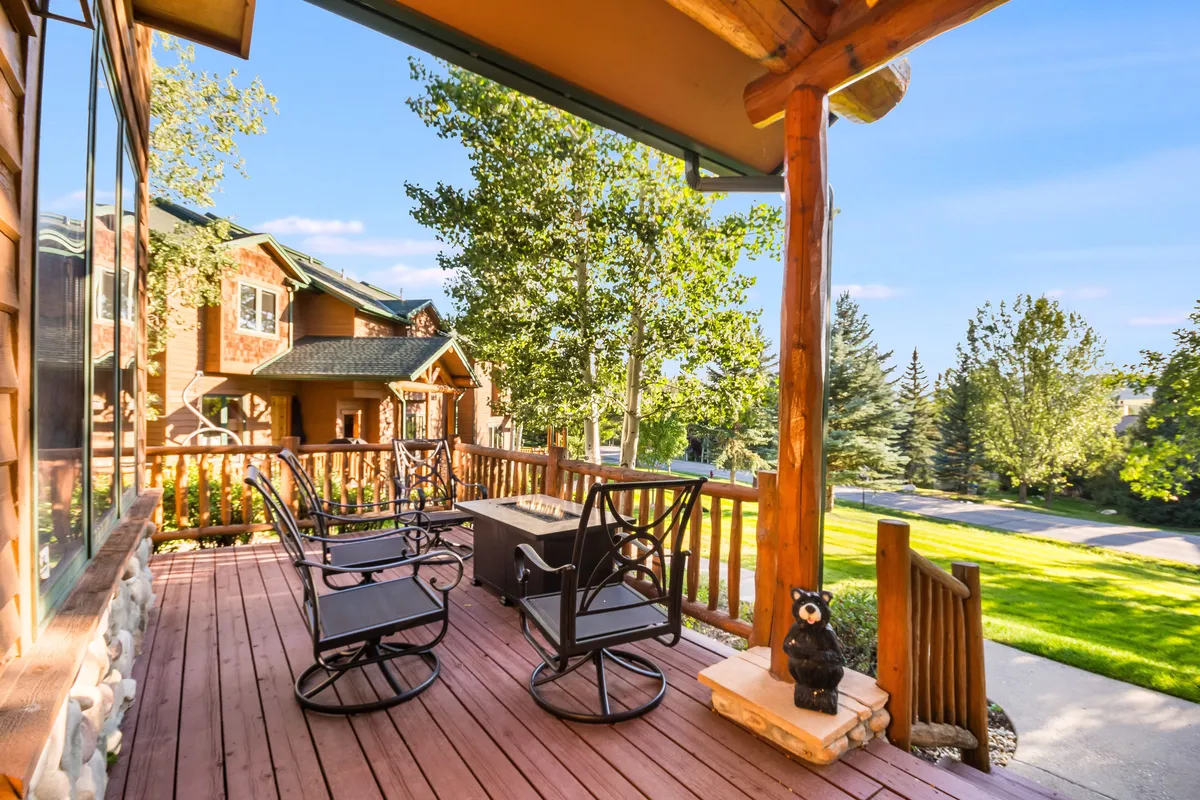 A rustic wooden deck set amongst trees features cozy chairs, a small table, and a whimsical bear sculpture. The tranquil setting emphasizes the peacefulness and connection to nature, offering a space for sunset viewing or socializing in the fresh air.