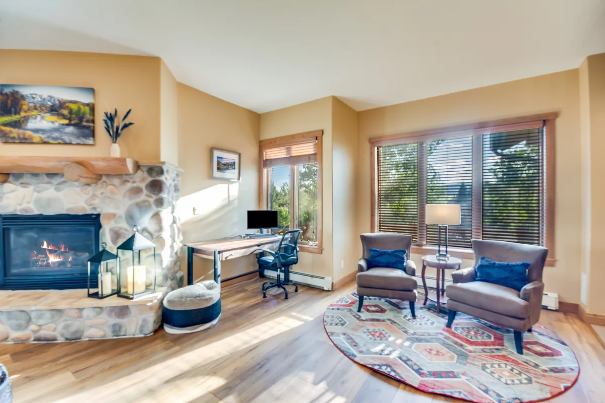 A bright, well-appointed living space with a fireplace, comfortable armchairs, a desk/workspace, a stylish rug, and large windows bringing in natural daylight. The room strikes a balance between relaxation and productivity, perfect for both leisure and high-speed wireless internet connectivity.