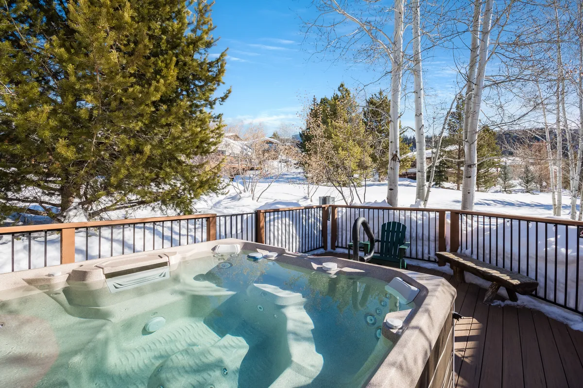 An exterior deck scene featuring a steaming hot tub surrounded by a snow-covered landscape, with a deck railing and a comfortable green chair. The backdrop creates a serene, inviting setting for relaxation after outdoor activities.