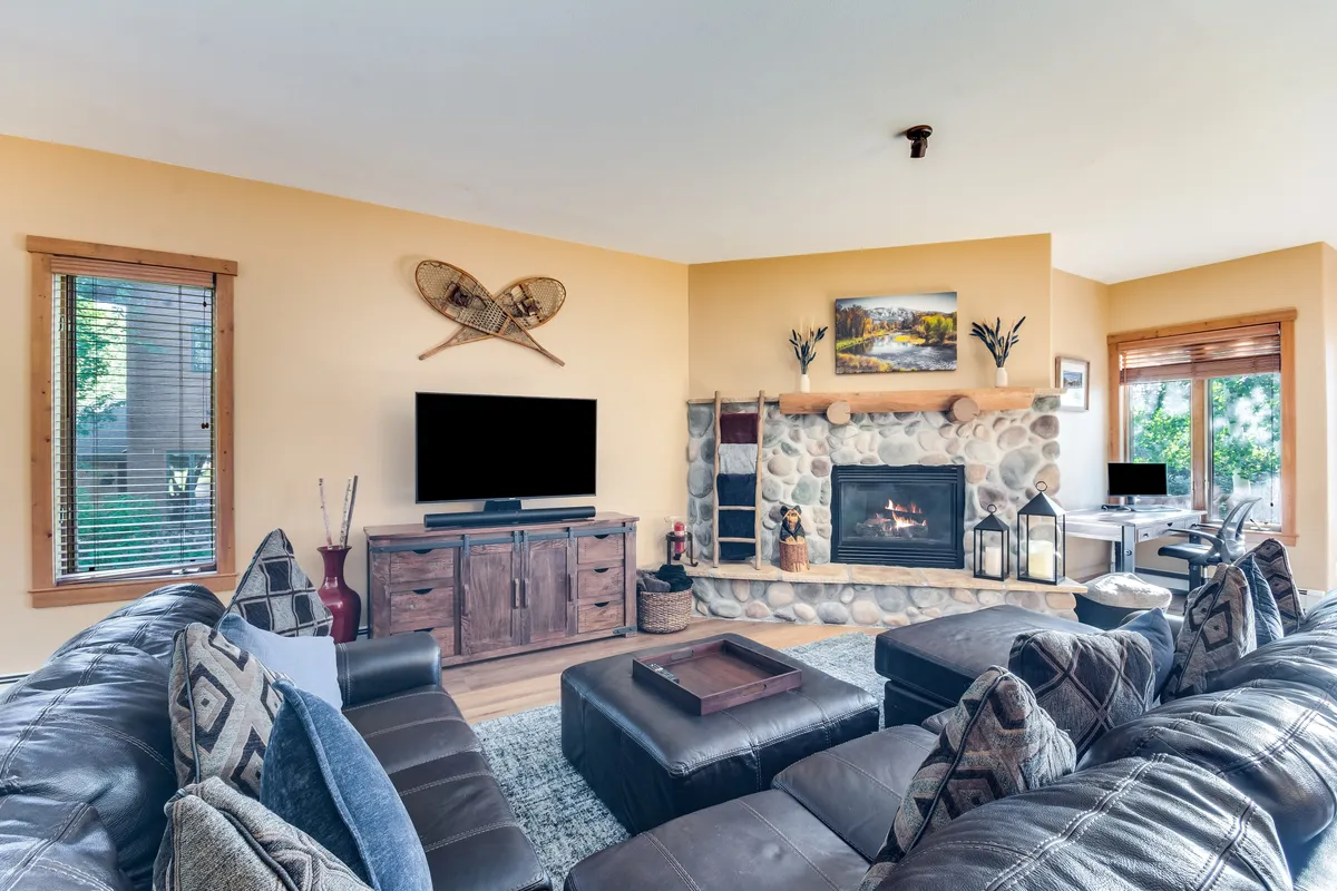 A spacious, open living room featuring a prominent stone fireplace, a large sectional sofa perfectly arranged for gatherings, a flat-screen television, and rich wooden furniture. Sunlight pours in from surrounding windows, illuminating the hardwood floors and creating a bright, welcoming ambiance.