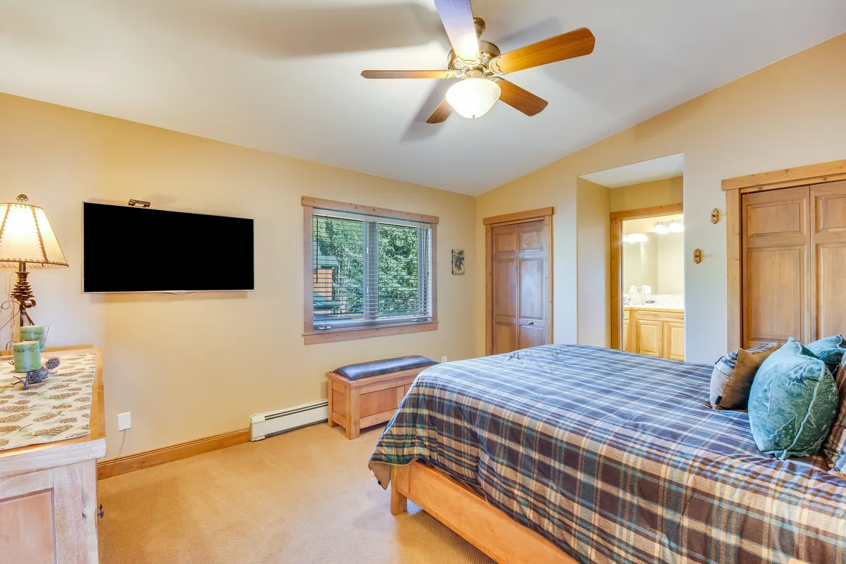 This image highlights a bedroom with a comfortable plaid bed, inviting lampshade, a window offering views outside, a flat-screen TV, and a wooden dresser. The overall atmosphere is cozy and restful, ideal for relaxation.